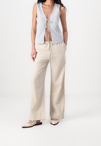 Gestreiftes ärmelloses Oberteil mit vorderem Bindeband, kombiniert mit beigen, weit geschnittenen Hosen und hellen Slippern. Weicher Stoff, lässige Passform.