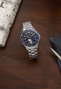 Ασημένιο ρολόι χειρός Timex με μπλε καντράν που φέρει σχέδιο Snoopy, τοποθετημένο σε ξύλινη επιφάνεια με κόμικ και στυλό κοντά.