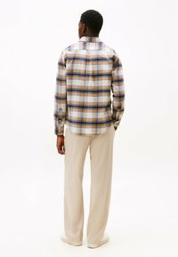 Button-up skjorte i ternet mønster med blå, gul og cremede farver. Lange ærmer, åben krave og buet kant. Båret med lysebeige bukser.