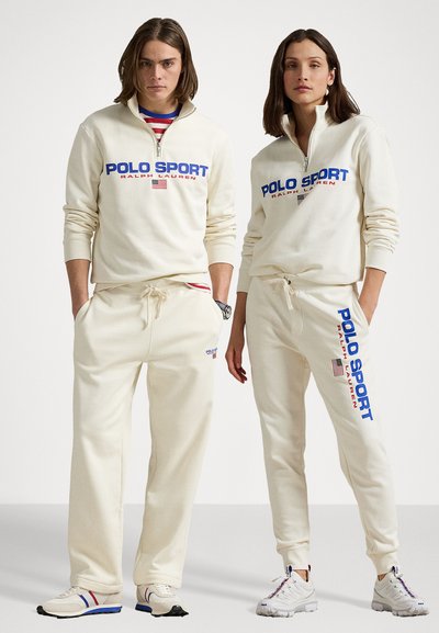 Set de îmbrăcăminte sport de culoare crem, compus dintr-o bluză cu fermoar și pantaloni jogger. Inscripții îndrăznețe în albastru "POLO SPORT" și accente cu steagul american. Textură moale.