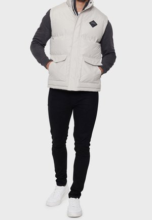 Chaleco puffy gris claro con un cuello alto, que cuenta con dos bolsillos frontales y un parche del logo en forma de diamante, usado sobre un suéter oscuro y pantalones negros.