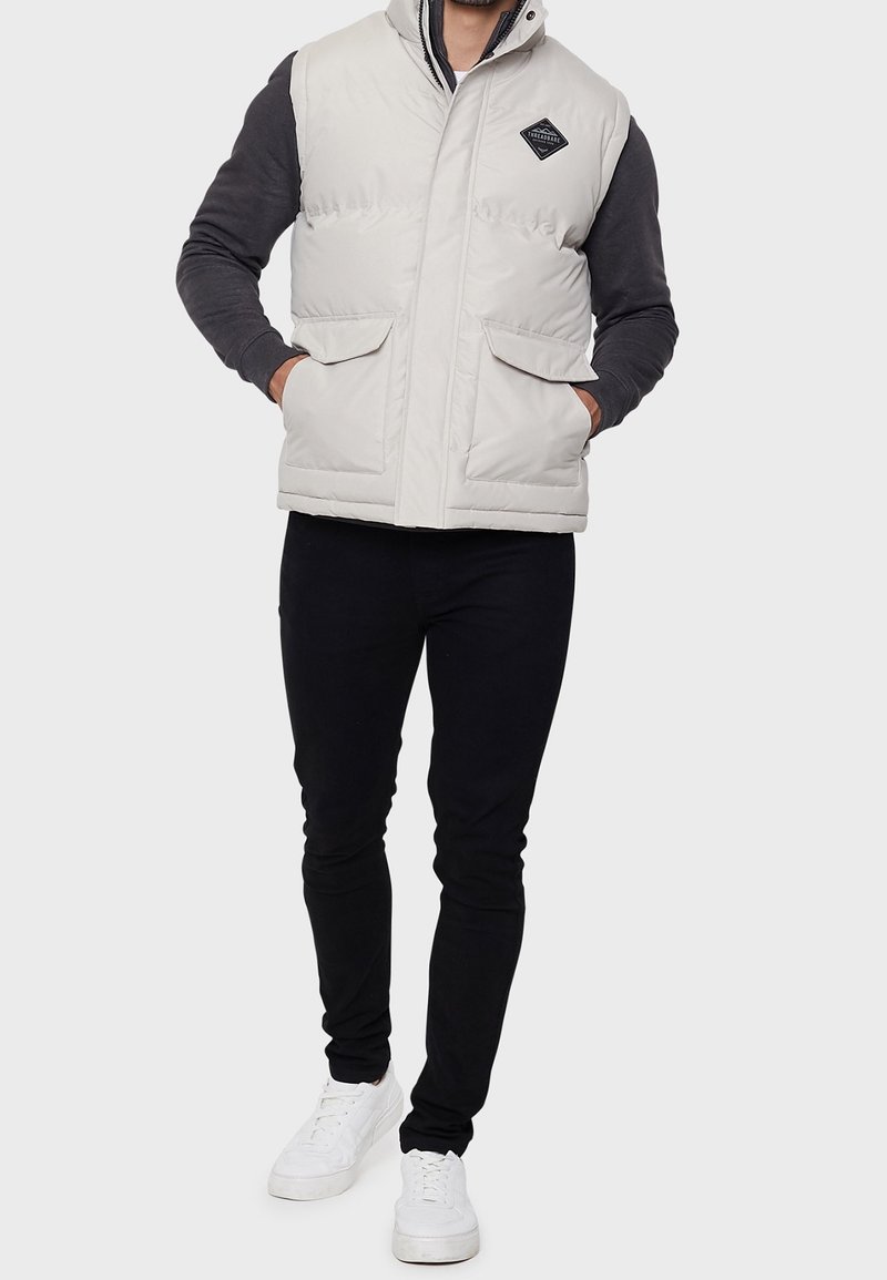 Helle graue Steppweste mit hohem Kragen, ausgestattet mit zwei Fronttaschen und einem Diamant-Logo-Patch, getragen über einem dunklen Pullover und schwarzen Hosen.