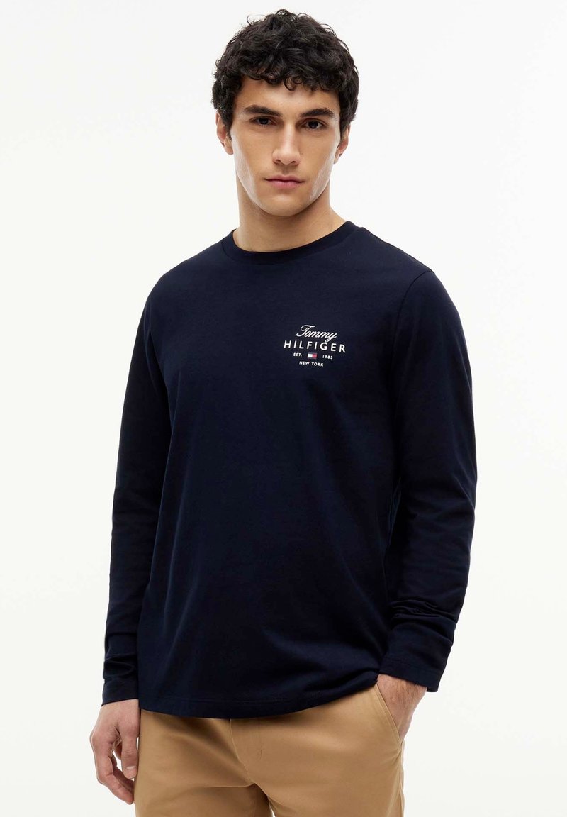 Jeune homme aux cheveux bouclés et foncés portant une chemise Tommy Hilfiger bleu marine à manches longues et un pantalon beige sur fond blanc.