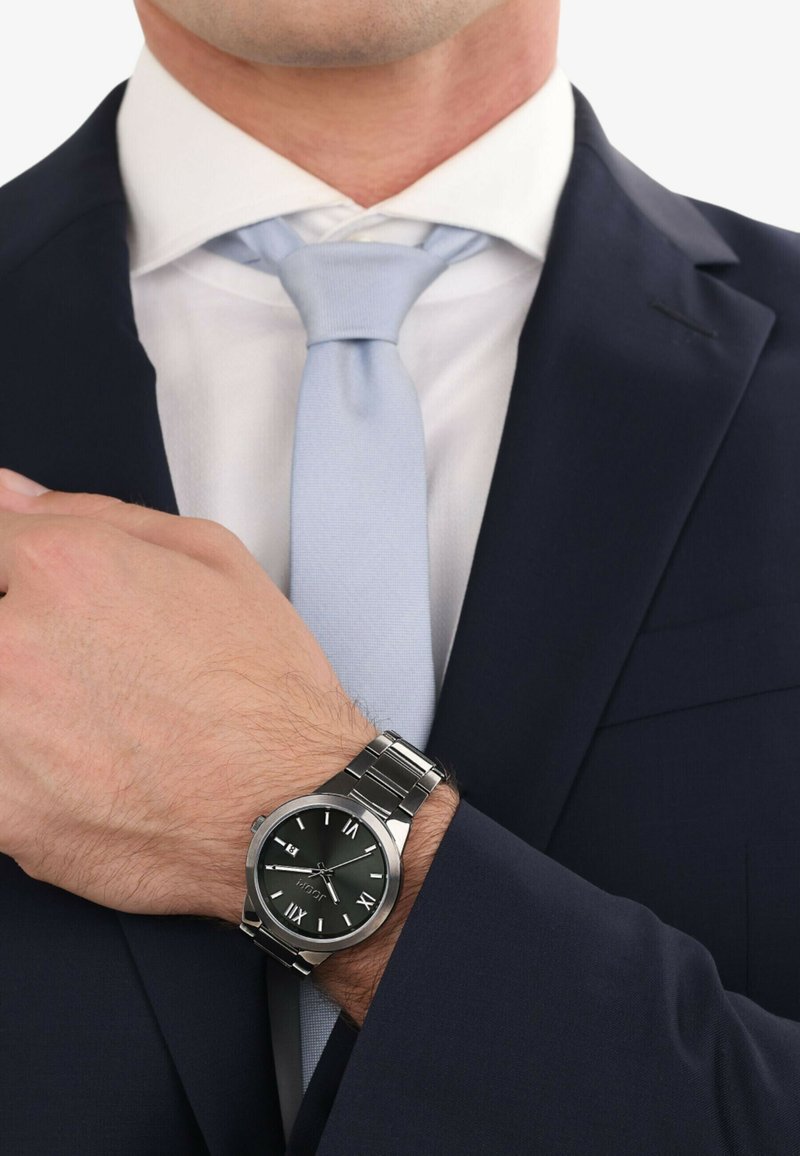 Silberne Metall-Uhr mit einem runden schwarzen Zifferblatt, römischen Ziffern und einem Armband aus Edelstahl. Am Handgelenk unter einem dunklen Anzug getragen. Hellblaue Krawatte.