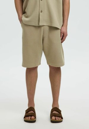 Homme portant un short beige texturé et une chemise boutonnée assortie, associé à des sandales ouvertes marron, debout devant un fond uni.