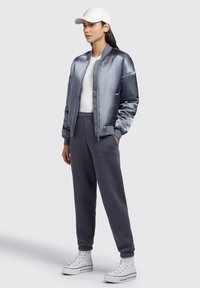 Jachetă bomber din satin de culoare gri deschis, tricou alb, pantaloni de trening gri închis și teniși albi cu guler înalt. Are un fermoar frontal și manșete ribetite.