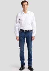 Egy férfi áll szemben, fehér gombos ingben, kék farmerben, fekete övvel és fekete csizmában, egyszínű világosszürke háttér előtt.