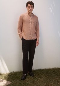 Camicia a maniche lunghe con bottoni di colore arancione chiaro con motivo geometrico, abbinata a pantaloni neri e scarpe nere intrecciate, sullo sfondo di un muro bianco.