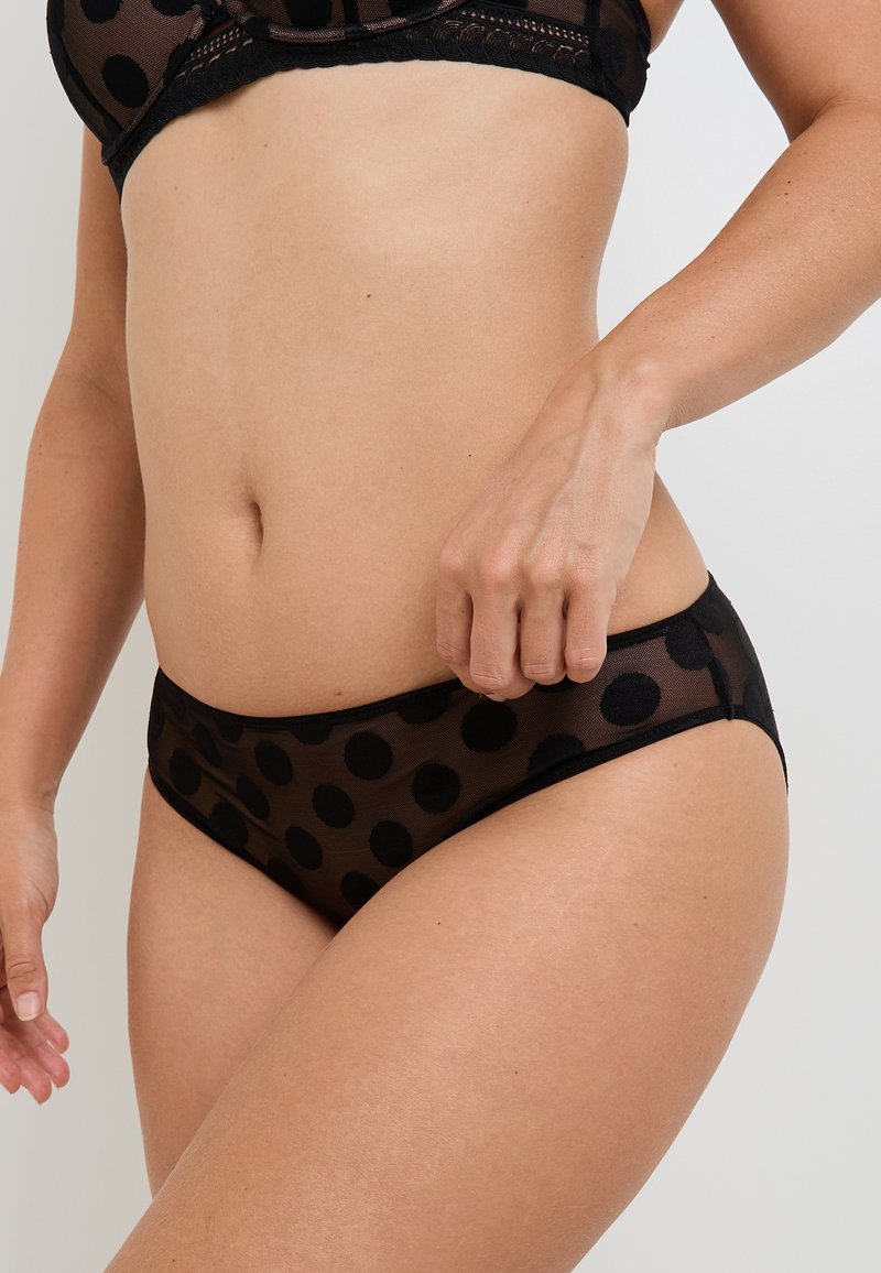 Donna che indossa lingerie a rete nera con grandi pois, che mostra il ventre e aggiusta l'elastico su uno sfondo semplice.