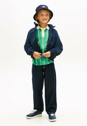 Dziecko w granatowym kapeluszu typu bucket, zielono-pasiastym polo, granatowej rozpinanej kurtce, ciemnych dżinsach i granatowych trampkach, stoi z rękami poprawiającymi kurtkę.