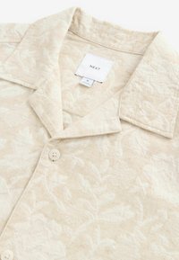 Camisa de manga corta color beige claro con un patrón floral texturizado, que presenta un cuello tipo solapa y dos botones claros. Etiqueta visible en el interior.