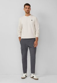 Licht beige sweatshirt met ribbelsteek in de hals, gecombineerd met donkergrijze cargo-broek met zijzakken, en witte sneakers met zwarte accenten.