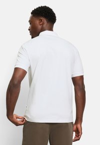 Weißes Poloshirt mit kurzen Ärmeln, geripptem Kragen und seitlichen Schlitzen. Glatte Textur, tailliertes Design und dezentes Branding am Saum.