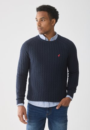 Pull en maille torsadée bleu marine avec un col rond et des poignets côtelés, superposé à une chemise à col bleu clair. Présente un petit logo rouge sur la poitrine.