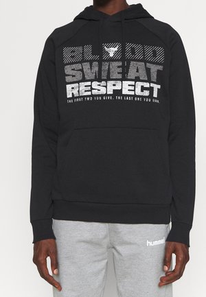 Person iført en sort hættetrøje med teksten "Blood Sweat Respect" og logo med en tyr, kombineret med lysegrå hummel træningsbukser.