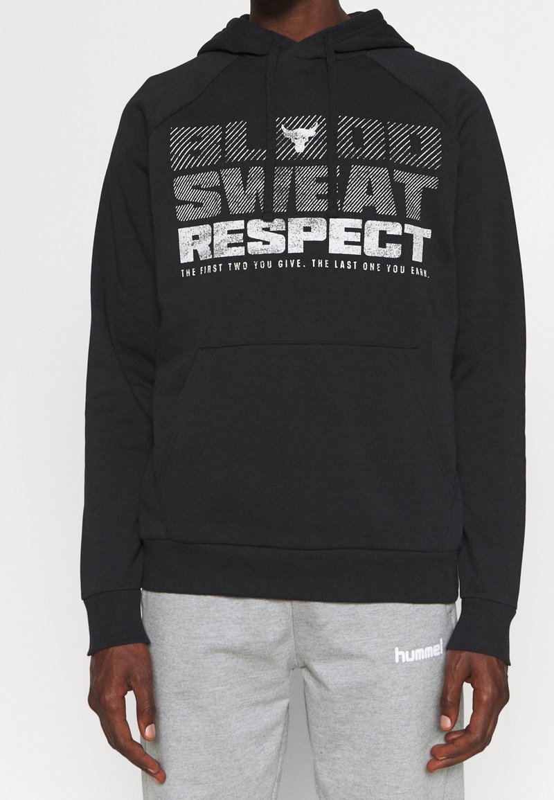 Personne portant un sweat à capuche noir avec le texte « Blood Sweat Respect » et un logo de taureau, assorti à un pantalon de jogging hummel gris clair.