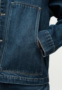 Τζιν μπουφάν με σκουρόχρωμο μπλε ύφασμα, έντονες ραφές, μεγάλη τσέπη και ελαφρώς υφή επιφάνειας. Καθαρός σχεδιασμός με εφαρμοστή γραμμή.