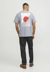 Pelēka T-krekls ar ģeometrisku dizainu, kurā ir sarkans aplis, baltas un pelēkas līnijas aizmugurē. Apvienots ar tumšiem denim džinsiem un brūnām kurpēm.