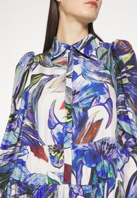 Femme portant une robe à manches longues avec un imprimé floral abstrait coloré et un col pointu, vue du cou à la taille.