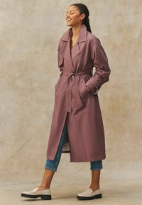 Hosszú, mauve színű trench coat, övvel a deréknál, széles hajtókákkal és oldalzsebekkel, világoskék cropped farmernadrággal és bézs loaferekkel párosítva.