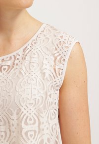 Ljus beige ärmlös topp i genomskinlig spets med intrikata blommönster, avslutad med en slät halsringning och kantade ärmhål.