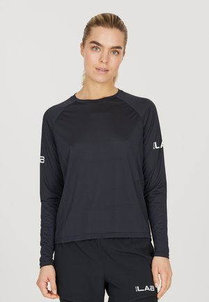 Pitkähihaine urheilu paita mustana, valmistettu kevyestä, sileästä kankaasta. Siinä on raglan-hihat ja logo-tyyppinen yksityiskohta vasemmassa hihassa.