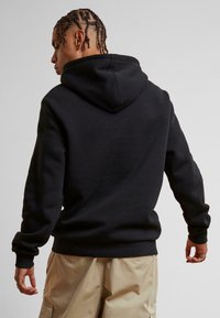 Schwarzer Kapuzenpullover mit strukturiertem Finish, ausgestattet mit Kapuze mit Kordelzug und gerippten Bündchen. Trägt khakifarbene Shorts und zeigt die Rückenansicht.