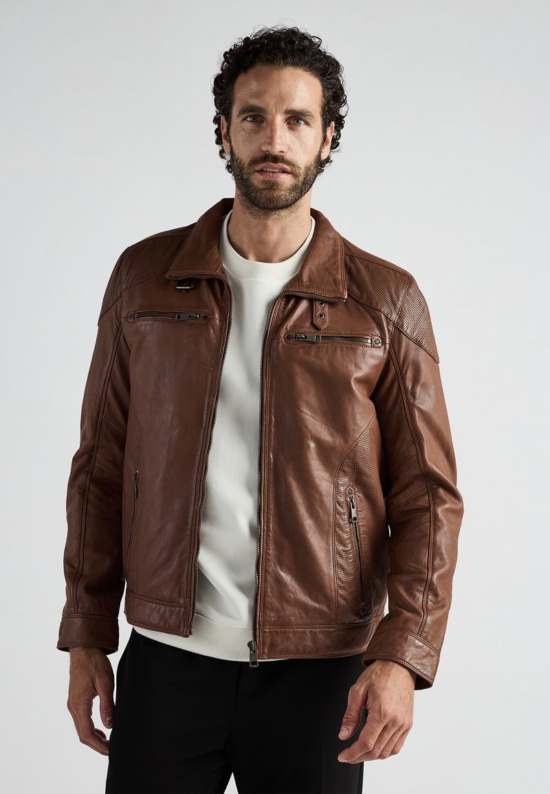 Veste en cuir marron avec un détail matelassé sur les épaules, des poches zippées et une finition lisse ; portée par-dessus une chemise de couleur claire.