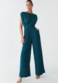 Petroleumsblå heldragt med vide ben, plisseret design og korte pufærmer. Har en figursyet talje og sidelommer. Blød stoftekstur.