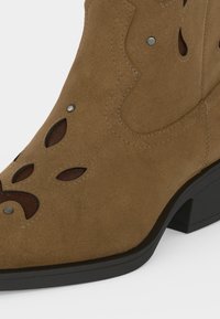 Bottine en daim marron avec motifs floraux ajourés, clous métalliques et petit talon bloc noir.