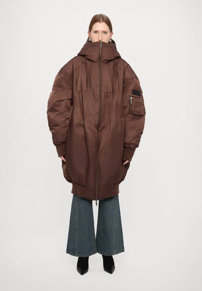 Giacca a vento oversized marrone con colletto alto, zip frontale e grandi tasche. Indossata sopra jeans di denim a gamba larga e scarpe nere.