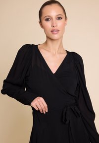 Sort omslagkjole med lange, puffete ermer, V-hals og knytedetalj i livet. Laget av lettvektig stoff med en glatt tekstur.