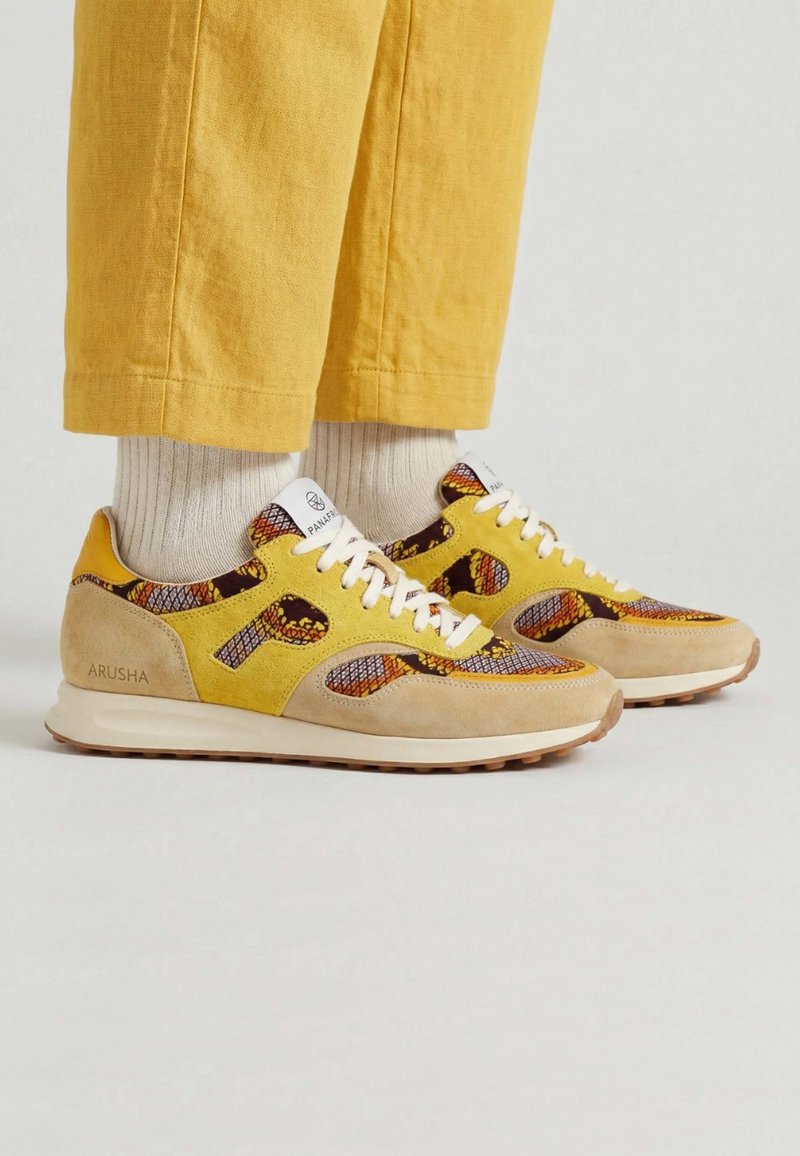 Des baskets jaunes et beiges avec un tissu à motifs multicolores, portées avec des chaussettes blanches et un pantalon jaune sur un fond clair uni.