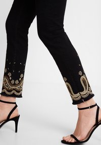 Piernas que llevan pantalones negros con detalles florales bordados en dorado cerca del dobladillo, combinados con sandalias negras de tacón alto con tiras al tobillo.