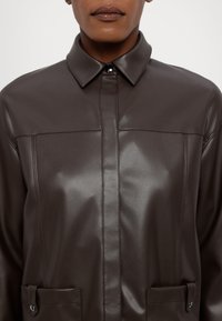 Gros plan sur une personne portant une veste en cuir brun foncé avec un col boutonné et des poches avant, sur un fond uni.
