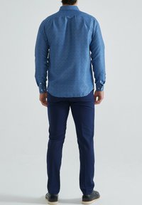 Chemise à manches longues bleue à motifs avec un col boutonné, assortie à un pantalon marine et des chaussures noires. Le tissu semble lisse et léger.