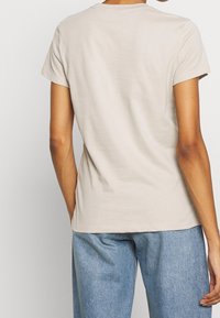 Camiseta de algodón color beige con mangas cortas, escote redondo y un ajuste relajado, combinada con pantalones de mezclilla azul claro. La vista trasera muestra una textura suave.