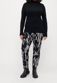 Osoba v černém roláku, černobílých kalhotách s abstraktním vzorem a černých kotníkových botách, stojící proti jednobarevnému pozadí.