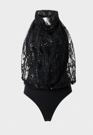 Femme aux longs cheveux foncés portant une combinaison noire sans manches à col halter avec des franges perlées et des talons ouverts noirs.