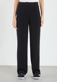 Pantalons larges noirs en tissu doux, avec une taille plate, ceinture cousue et poches latérales. Associés à des baskets sombres.