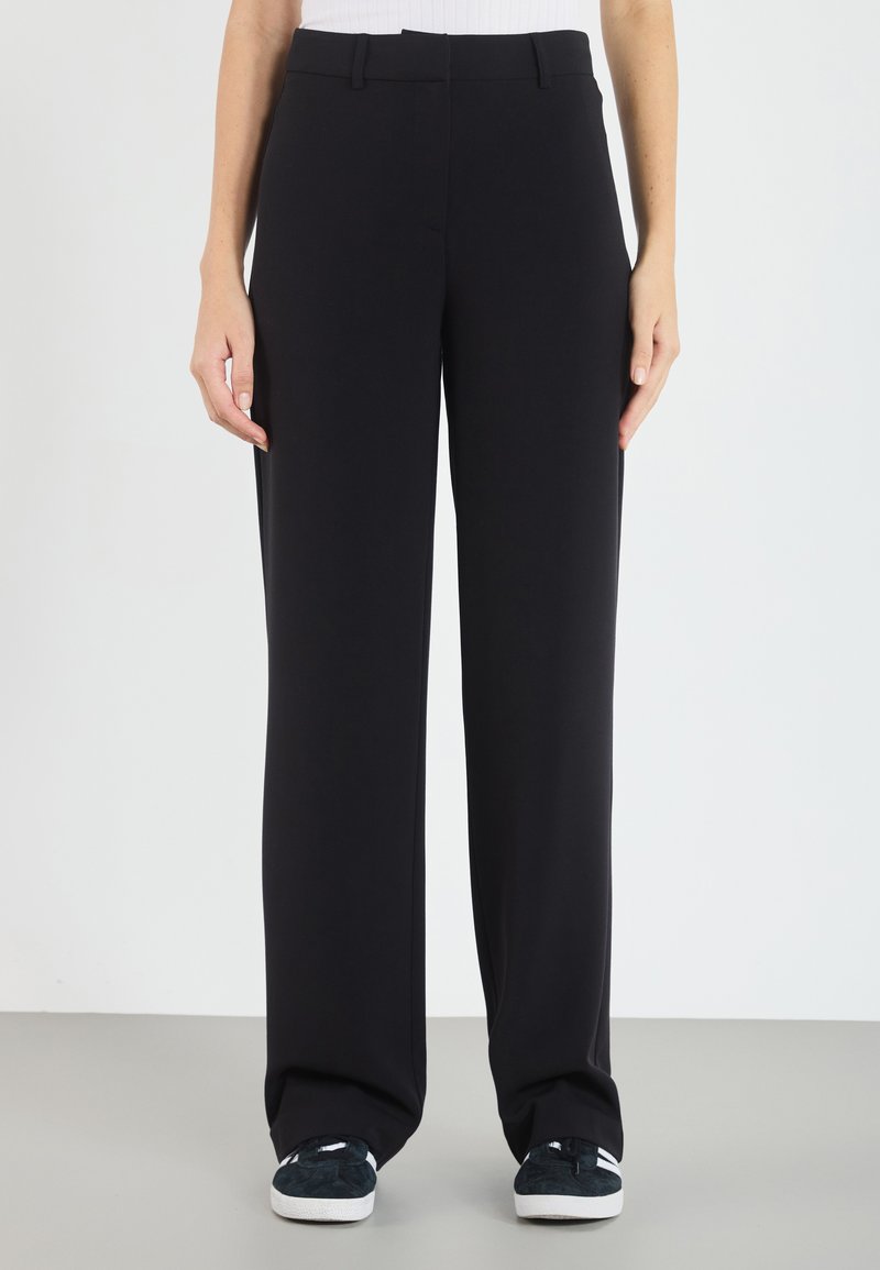 Pantalons larges noirs en tissu doux, avec une taille plate, ceinture cousue et poches latérales. Associés à des baskets sombres.