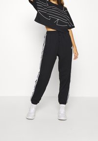 Svart kortärmad t-shirt med vita geometriska ränder, parat med svarta joggers med vita sidostripes och en broderad logotyp. Vita sneakers.