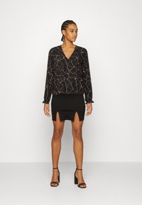 Blusa preta de mangas compridas com padrão abstrato em dourado, combinada com uma saia mini preta com duas fendas laterais. Modelo usa sandálias de salto pretas.