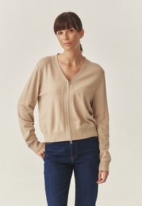 Beige kötött kardigán V-nyakkal, elöl cipzárral és bordázott szegéllyel. Hosszú ujjú, sima textúrájú, sötétkék farmernadrággal párosítva.