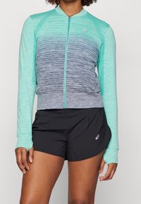 Letvægts, lynlåsjakke i gradient aqua og grå stof, med lange ærmer og en tætsiddende pasform. Sammen med sorte shorts.