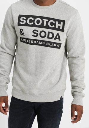 Mand iført en lysegrå sweatshirt med sort og hvid tekst "Scotch & Soda Amsterdams Blauw" og mørke bukser, stående med afslappede arme.
