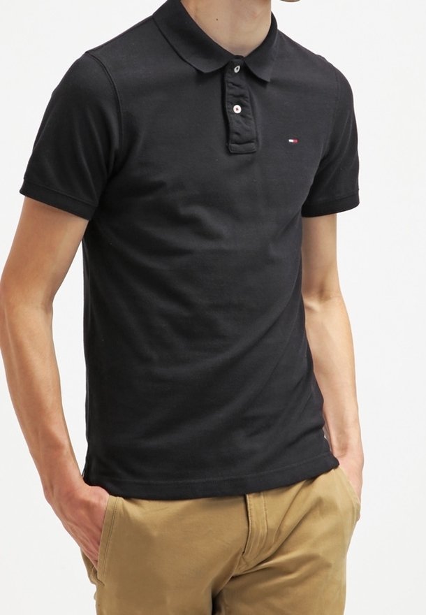 Homme portant un polo noir à manches courtes avec un petit logo sur la poitrine et un pantalon kaki, mains dans les poches, debout devant un fond blanc.