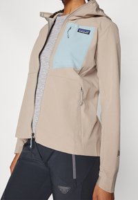 Nainen, jolla on vaaleanbeige huppari, jossa on vaaleansininen vetoketjullinen rintatasku, ja mustat urheiluhousut, joissa on logo reidessä.