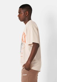 Beige bomullströja med en orange grafisk tryck, avslappnad passform, korta ärmar och rundad halsringning, bärs med bruna joggingsbyxor.