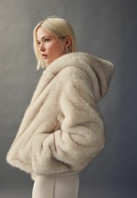 Blondína v béžovej kabátovej bunde z umelej kožušiny s kapucňou, so zdvihnutými rukami v vreckách, otočená k ľavej strane na jednotnom šedom pozadí.